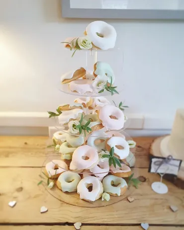 Acrylic tower stacked with pastel pink, mint and white glazed donuts decorated with fresh flowers on a wooden table